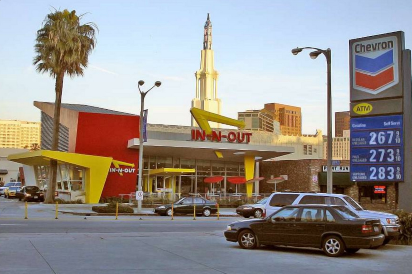 In-N-Out Burger Restaurant in California dominates the center of the frame, with a palm tree and a tower, wide roads, several cars, and no humans in sight in late afternoon. A Chevron gas station sign is in front and to the side. Multiple holders for broken flex-posts are in the street.