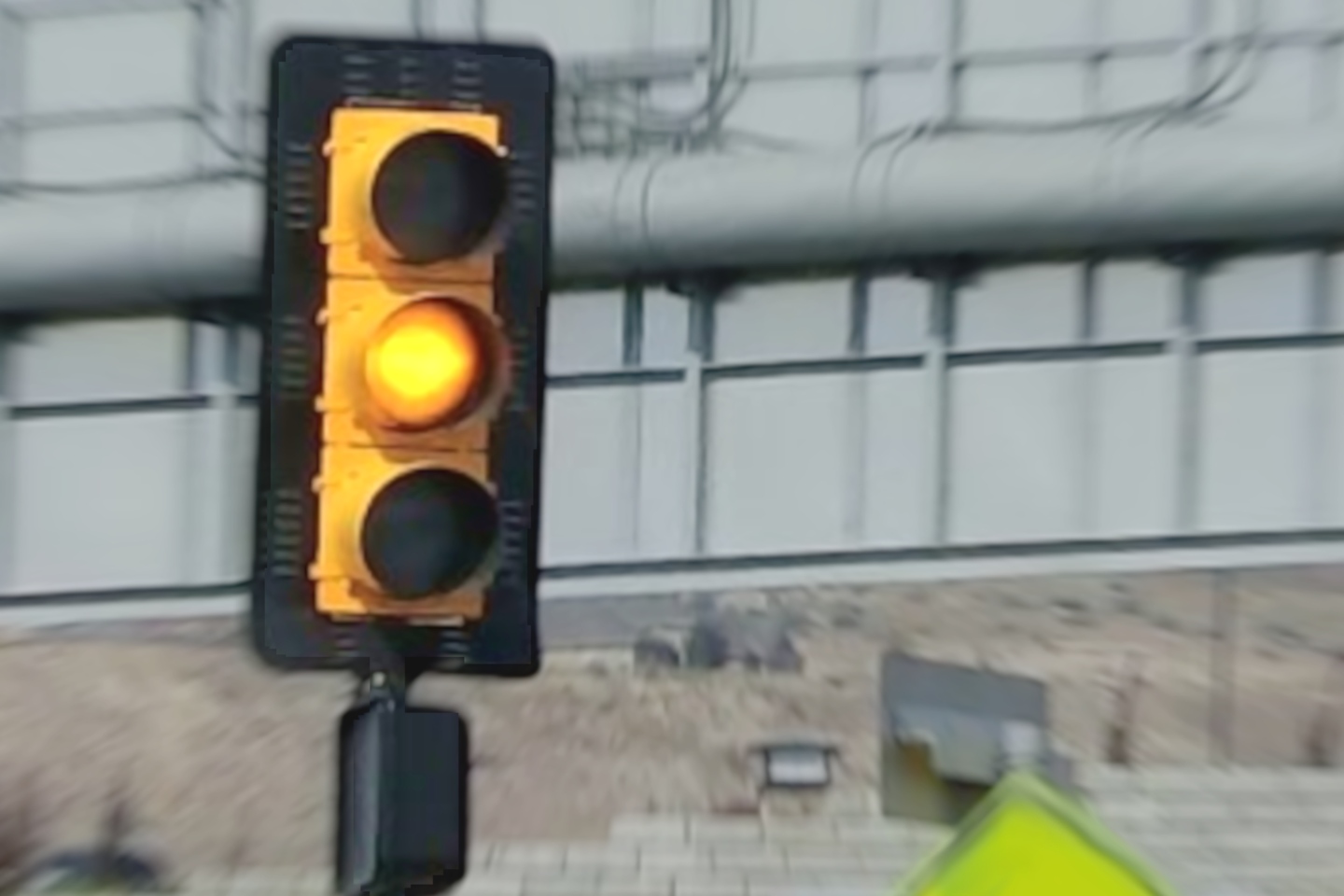 A vertical traffic light on a post is on the yellow part of the light cycle. Behind are the back of a highway sign, a diamond crossing sign, and a concrete wall to hold back dirt and sand.