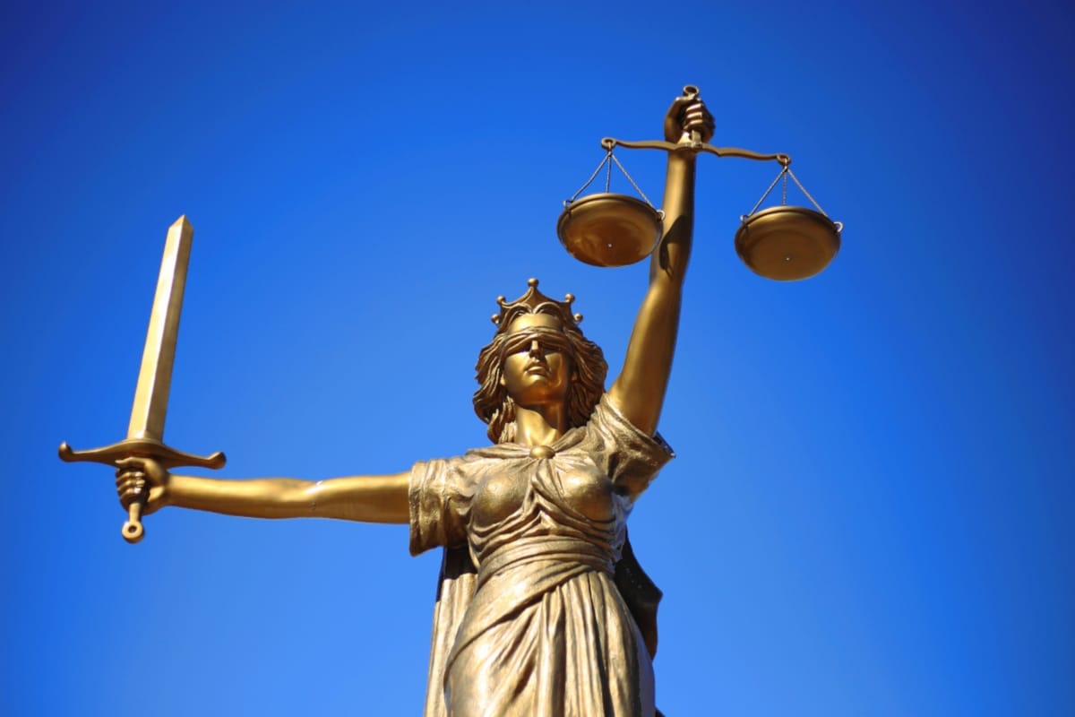 Against a bright blue sky, a golden statue of blinded Lady Justice holds up scales in one and a sword in the other.